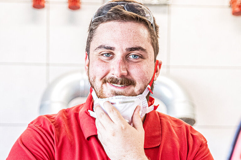 Ouvrier en combinaison rouge et casque devant un boîtier électrique. Il pince un câble avec une pince.