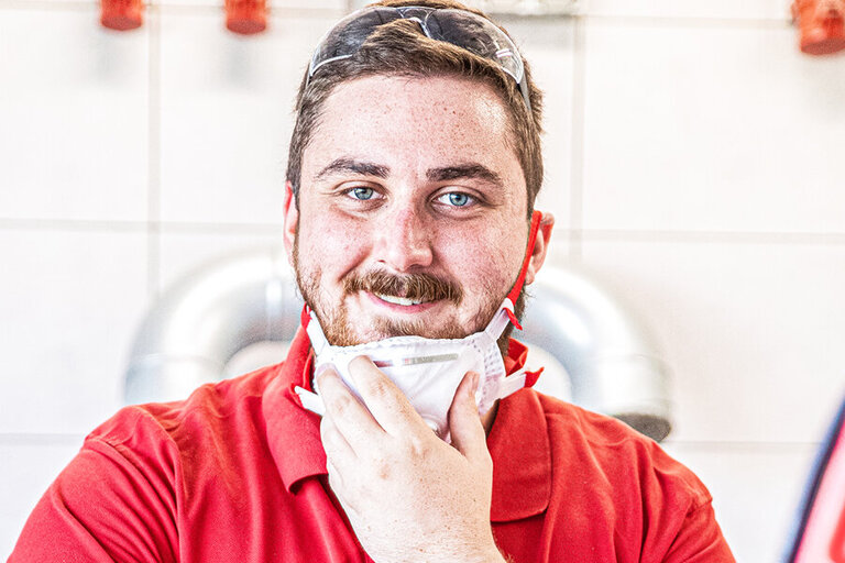 Arbeiter mit rotem Shirt lächelt in die Kamera und hat die Staubschutzmaske heruntergezogen.
