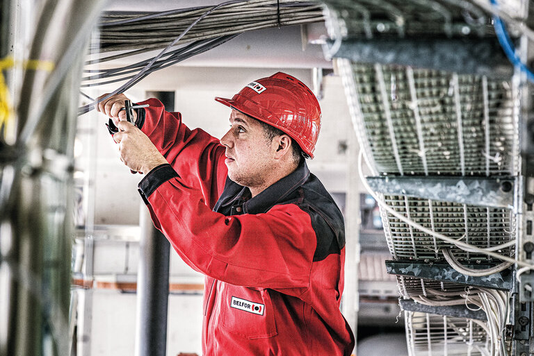 Arbeiter in rotem Overall und mit Helm an einem Schaltkasten. Er kneift mit der Zange ein Kabel.