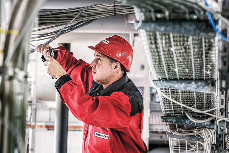 Arbeiter in rotem Overall und mit Helm an einem Schaltkasten. Er kneift mit der Zange ein Kabel.