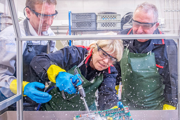 Drei Personen in Schutzbrillen, Schürzen und Handschuhen arbeiten gemeinsam an einer elektronischen Leiterplatte.