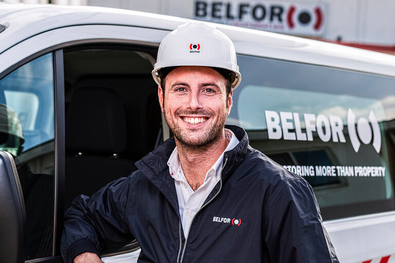 BELFOR-medewerker met helm staat glimlachend voor een bedrijfswagen.