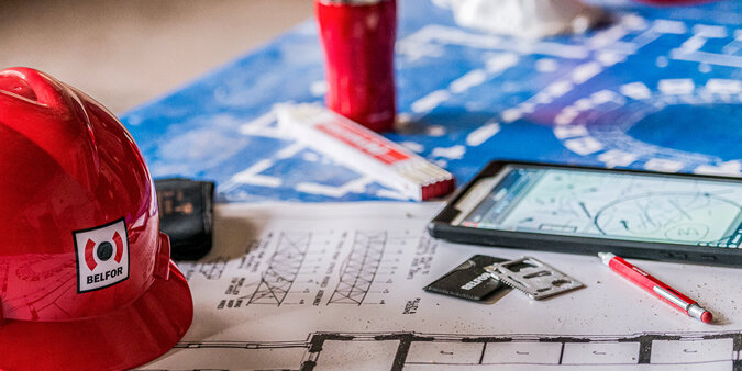 belfor_bewerbung Arbeitsplatz mit Bauhelm, Grundriss, Stift und Kaffeebecher